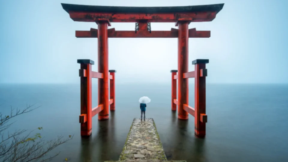 Vermilion torii gate standing in mist on Lake Ashi at Hakone during Japan's tsuyu rainy season, with a visitor holding an umbrella on the stone pier