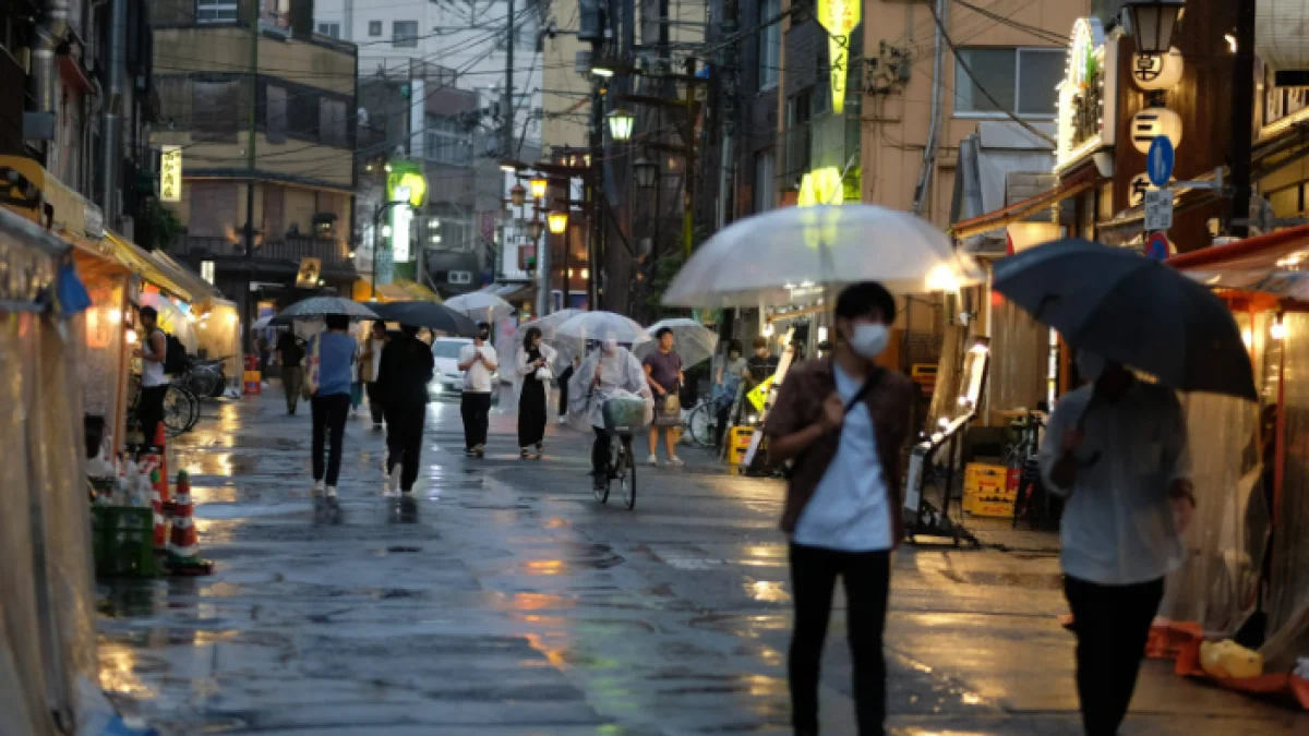 Pedestrians with umbrellas on a rain-slicked Tokyo street during tsuyu, with shop lights reflecting on wet pavement